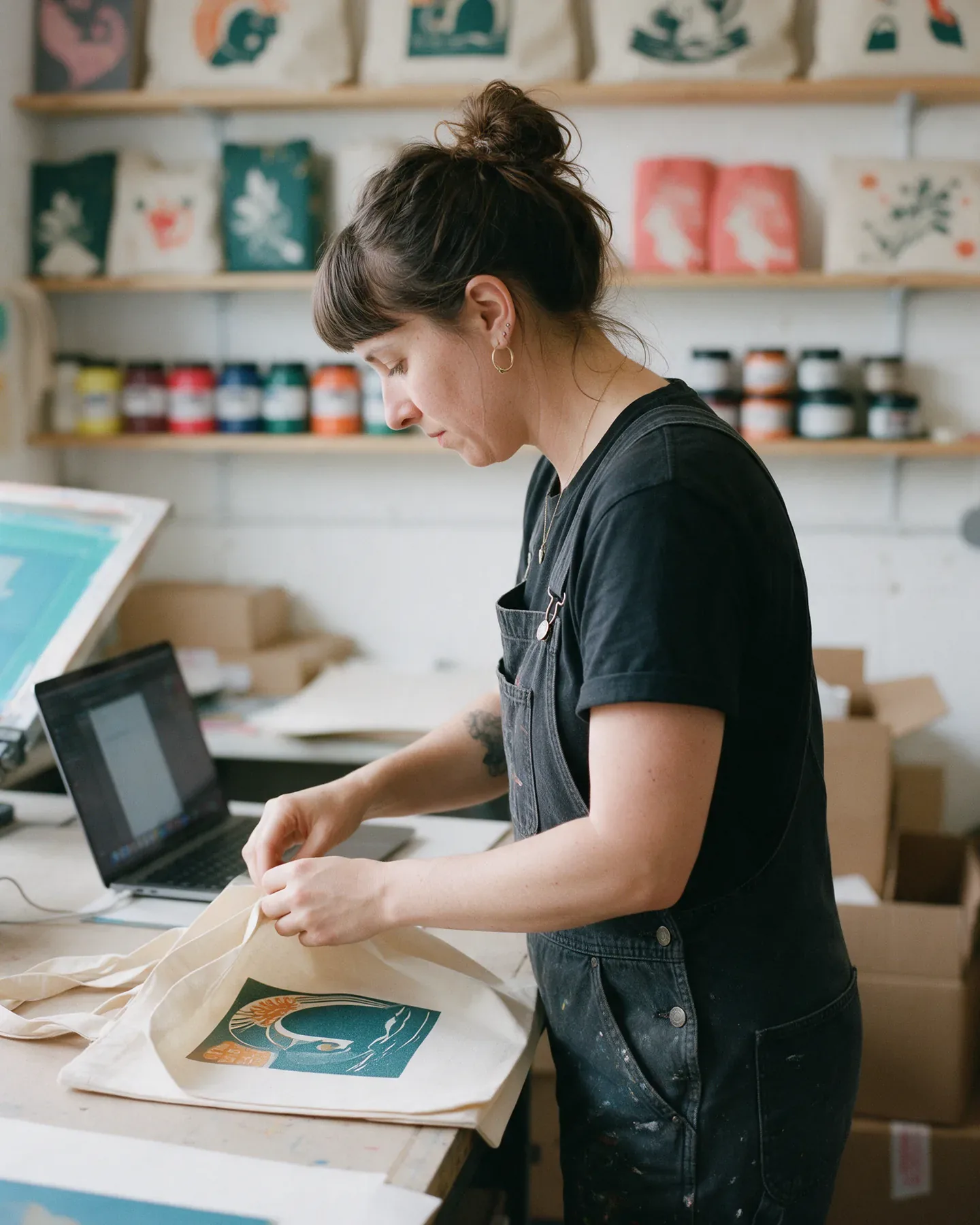A maker folding a printed tote bag beside her laptop in a screen-printing studio.
